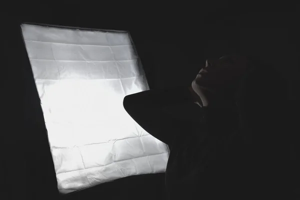 Tanımlanamayan genç bir kadın saçını düzeltiyor ve karanlık odada parlayan lambanın yanına bakıyor. 