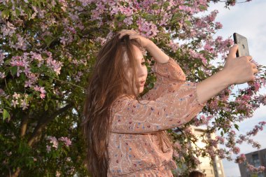 Gün batımında çiçek açan ağacın yanında dinlenirken akıllı telefondan selfie çeken genç ve odaklanmış Asyalı güzel bir kadın. 