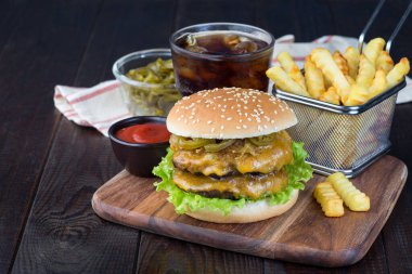 İki köfteli çizburger, çedar peyniri, marul, karamelize soğan ve jalapeno dilimleri, patates kızartması ve soda ile servis ediliyor, tahta tahta üzerinde, yatay, kopyalama alanı.