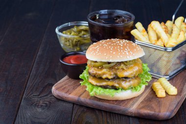 Çift peynirli burger, iki köfte, çedar peyniri, marul, karamelize soğan ve jalapeno dilimleri, patates kızartması ve soda ile servis edilmiş, tahta bir tahta üzerinde, yatay, kopyalama alanı.
