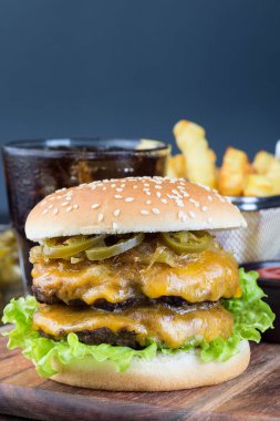 Duble peynirli burger, iki köfte, çedar peyniri, marul, karamelize soğan ve jalapeno dilimleri, patates kızartması ve soda ile servis edilmiş, tahta tahtada, dikey, kopyalama alanı.