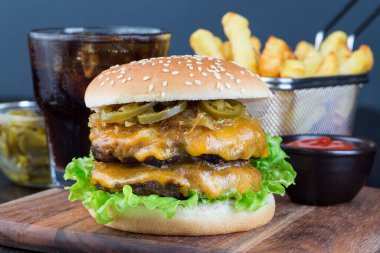 Çift peynirli burger, iki köfte, çedar peyniri, marul, karamelize soğan ve jalapeno dilimleri, patates kızartması ve soda ile servis edilir, tahta bir tahtanın üzerinde, yatay, yakın plan