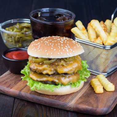 Çift peynirli burger, karamelli soğan, marul ve jalapeno, patates kızartması ve soda ile servis ediliyor, tahta bir tahtanın üzerinde, kare şeklinde.