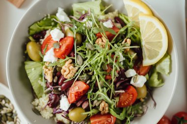 Sağlıklı taze sebze salatası, salatalık, domates, ıspanak, roka, zeytin ve peynir salatası beyaz bir masada, balkabağı, ceviz ve sarı limonlu ayçekirdeği. Yemek geçmişi, yemek tarifi. Yukarıdan yakından, çiğ gıda fikri