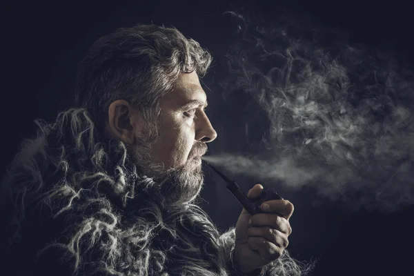 Solid bearded man in a fur mantle smoking pipe