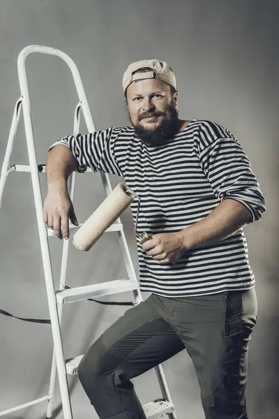 Vreugdevolle bebaarde ambachtsman met kwastroller en ladder — Stockfoto