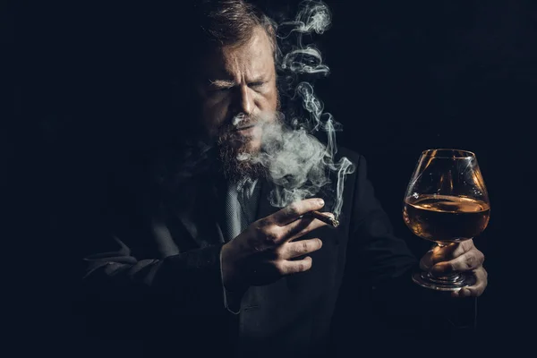 Solide homme barbu confiant en costume avec verre de whisky et cigare avec fumée — Photo