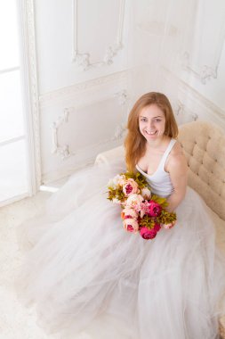 Çiçekleri olan, geniş etekli beyaz elbiseli güzel bir kadın kanepede oturuyor.