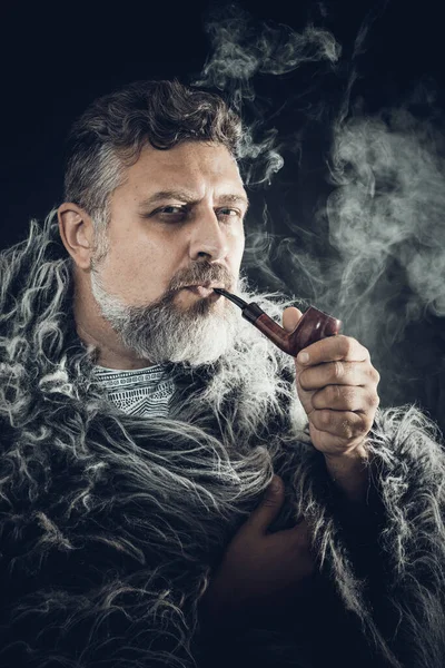 Solid bearded man in a fur mantle smoking pipe