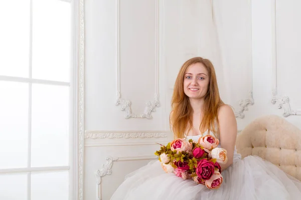 Gyönyörű fiatal nő, virágokkal, fehér ruhában, széles szoknyában, a kanapén ülve. — Stock Fotó