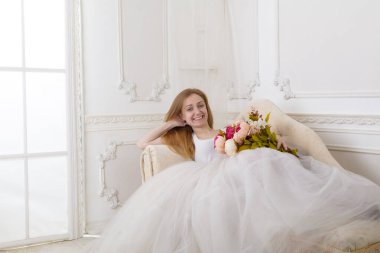 Çiçekleri olan, geniş etekli beyaz elbiseli güzel bir kadın kanepede oturuyor.