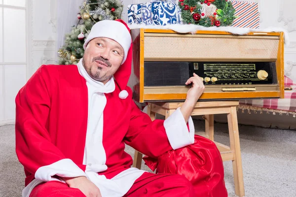 Γενειοφόρος άνδρας ντυμένος με κοστούμι Άγιος Βασίλης κάθεται κοντά στο ρετρό ραδιόφωνο που — Φωτογραφία Αρχείου