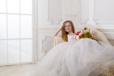 Çiçekleri olan, geniş etekli beyaz elbiseli güzel bir kadın kanepede oturuyor.