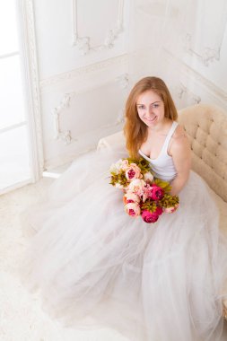 Çiçekleri olan, geniş etekli beyaz elbiseli güzel bir kadın kanepede oturuyor.