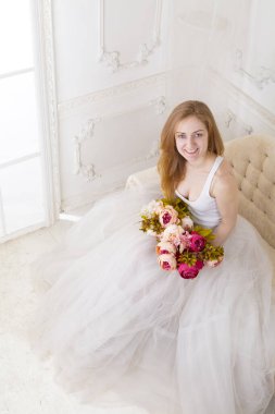 Çiçekleri olan, geniş etekli beyaz elbiseli güzel bir kadın kanepede oturuyor.