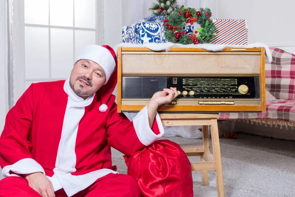 Γενειοφόρος άνδρας ντυμένος με κοστούμι Άγιος Βασίλης κάθεται κοντά στο ρετρό ραδιόφωνο που — Φωτογραφία Αρχείου