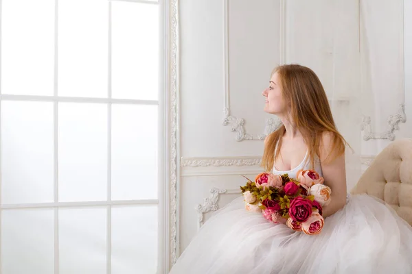 Mulher bonita com flores vestidas com um vestido branco cheio de rodapé largo sentado no sofá — Fotografia de Stock