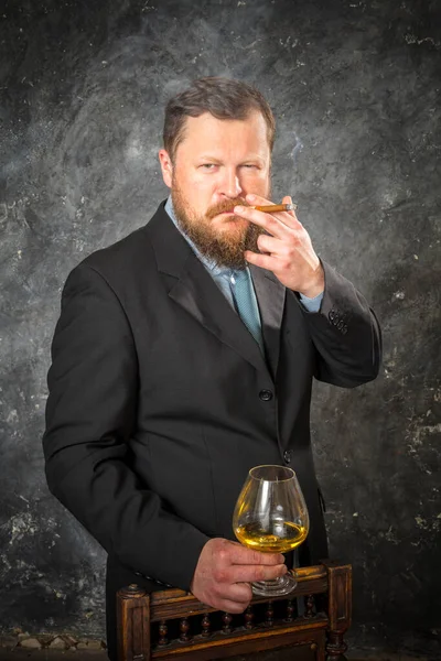 Stevige zelfverzekerde man met baard in pak met glas whisky en een ciga — Stockfoto