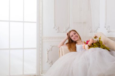 Çiçekleri olan, geniş etekli beyaz elbiseli güzel bir kadın kanepede oturuyor.