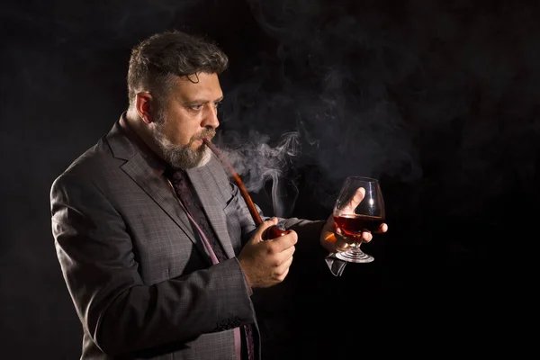 Hombre barbudo de confianza sólida en traje con vaso de whisky pipa humeante — Foto de Stock