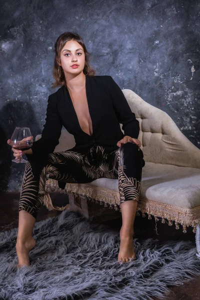 Piękna młoda dama w czarnej sukience siedzi na kanapie ze szklanką wina — Zdjęcie stockowe
