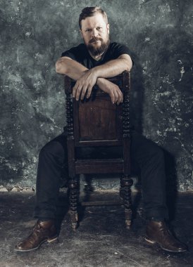 Tişört giymiş, sandalyede oturan olgun sakallı adam.