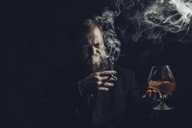 Bir bardak viski ve dumanlı puroyla takım elbiseli, kendinden emin, sakallı bir adam.