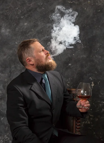 Stevige zelfverzekerde man met baard in pak met glas whisky en rook — Stockfoto