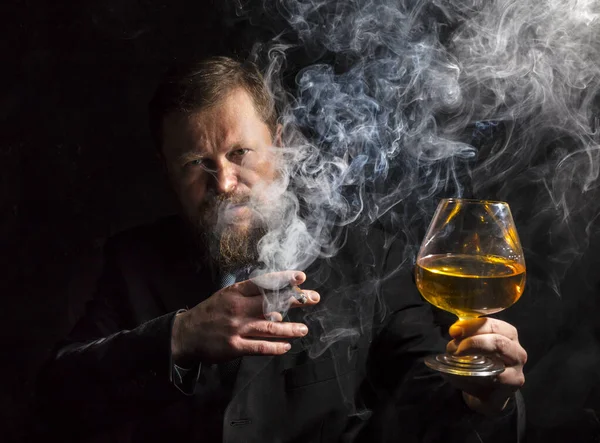 Hombre barbudo sólido y confiado en traje con vaso de whisky y cigarro con humo — Foto de Stock