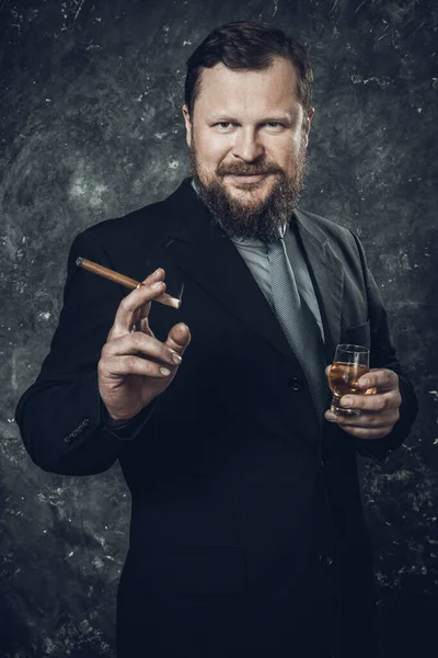 Solido uomo barbuto fiducioso in abito con bicchiere di whisky e sigaro — Foto Stock