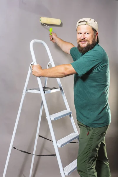 Capataz barbudo alegre em camiseta verde com rolo de escova e escada — Fotografia de Stock