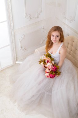 Çiçekleri olan, geniş etekli beyaz elbiseli güzel bir kadın kanepede oturuyor.