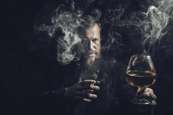 Fast säker skäggig man i kostym med glas whisky och cigarr med rök — Stockfoto