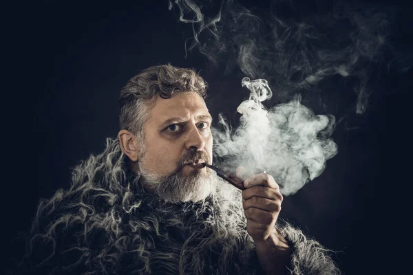 Hombre barbudo sólido en un manto de piel pipa humeante — Foto de Stock