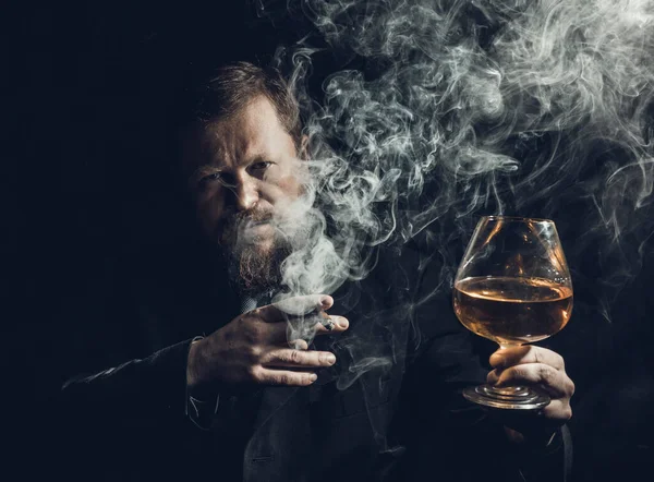 Solide homme barbu confiant en costume avec verre de whisky et cigare avec fumée — Photo