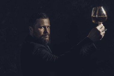 Bir bardak viskiyle takım elbiseli, kendinden emin, sakallı bir adam.