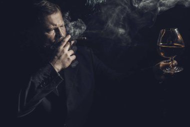 Bir bardak viski ve dumanlı puroyla takım elbiseli, kendinden emin, sakallı bir adam.