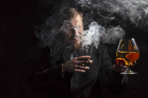 Pewny siebie brodaty mężczyzna w garniturze ze szklanką whisky i cygarem z oparami — Zdjęcie stockowe