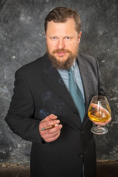 Solido uomo barbuto fiducioso in abito con bicchiere di whisky e sigaro — Foto Stock