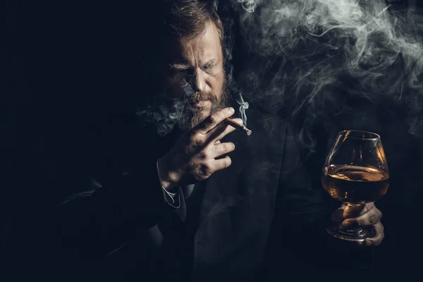Solide selbstbewusste bärtige Mann im Anzug mit Glas Whisky und Zigarre mit Rauch — Stockfoto