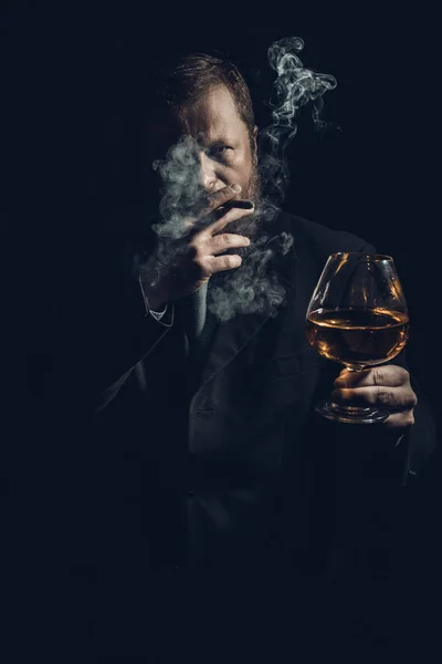 Pewny siebie brodaty mężczyzna w garniturze ze szklanką whisky i cygarem z oparami — Zdjęcie stockowe