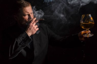 Bir bardak viski ve dumanlı puroyla takım elbiseli, kendinden emin, sakallı bir adam.