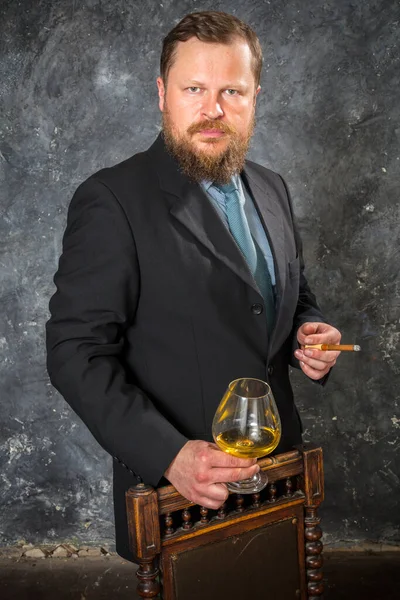 Solido uomo barbuto fiducioso in giacca e cravatta con un bicchiere di whisky e una sigaretta — Foto Stock