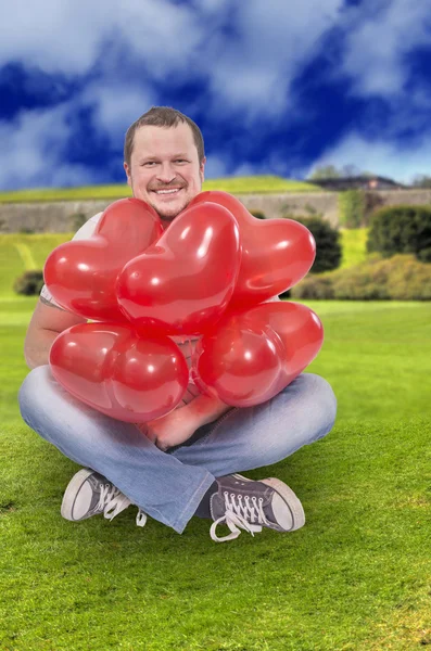 Mladý muž sedí s červenými balóny s zelený trávník pole v pozadí — Stock fotografie