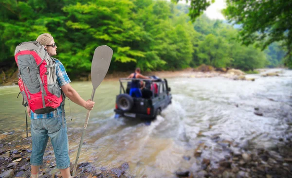 Tourist man with backpack and paddle watching the 4x4 vehicle crossing the river — Stock Photo, Image