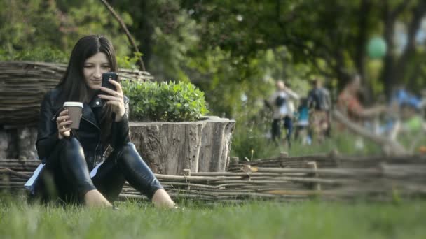 Μια νεαρή γυναίκα όμορφη, χρησιμοποιώντας μια κινητή εφαρμογή και πίνοντας τον καφέ στο πάρκο — Αρχείο Βίντεο