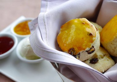 Çilek reçeli, marmelat ve kremayla eşleştirilmiş, kumaş kaplı bir sepsiyonda servis edilen taze pişmiş altın çörekleri yakından çek. Temiz, parlak ve iştah açıcı yemek tasarımı kafe menüleri, fırın promosyonları ve ticari reklamlar için idealdir. Yumuşak hamur işi