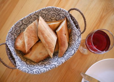 Tepeden aşağı ve yakın planda, hasır sepette servis edilen ve desenli kumaşla kaplanmış taze dilimlenmiş Fransız baget ekmeği. Kabuklu altın-kahverengi ekmek bir bardak kırmızı şarap veya meyve suyu ve beyaz bir tabakla birlikte açık ahşap bir masanın üzerine yerleştirilir.