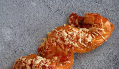 Erimiş peynir ve baharatlı sosla kaplanmış lezzetli bir poğaçanın yakın görüntüsü. Fırının altın-kahverengimsi yumuşacık dokusu, en az gri beton arka planıyla çok güzel zıtlaşır. Kahvaltı ya da atıştırmalık için mükemmeldir.