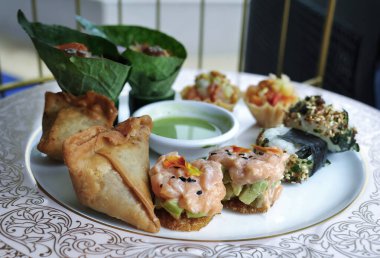 Dekoratif porselen tabakta servis edilen enfes bir meze tabağının yakından görüntüsü. Seçeneklerde kızarmış samosa, avokado ile taze somon tartar ve Miang Kham (betel yaprağı ambalajı) gibi geleneksel Tayland aperatifleri yer alıyor.